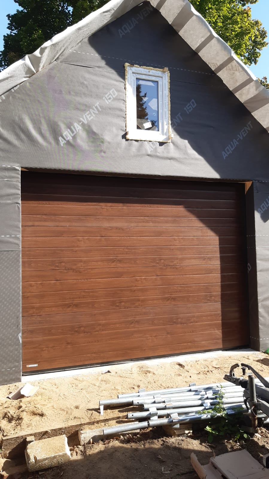 Nowa brama garażowa segmentowa w kolorze drewna, zamontowana w budynku w trakcie budowy, widoczne materiały izolacyjne i okno nad bramą.