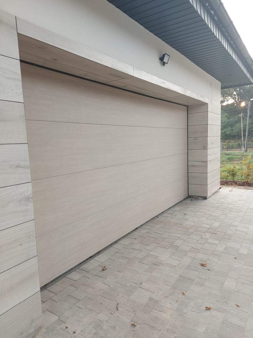 Zamknięta brama garażowa segmentowa w kolorze jasnego drewna, z poziomymi panelami, zamontowana w jasnym garażu z kostki brukowej na podjeździe.