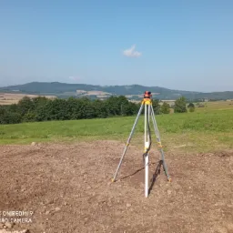 Geodezyjny niwelator optyczny ustawiony na trójnogu na przygotowanym pod budowę terenie, w tle zielone wzgórza i błękitne niebo z pojedynczą chmurą.