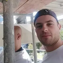 Portret młodego mężczyzny w czapce, uśmiechającego się do kamery na tle rusztowania i częściowo widocznej elewacji budynku w trakcie prac. W tle widoczny fragment innego pracownika i zabudowa miejska.