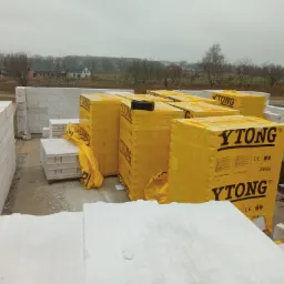 Budowa domu z bloczków Ytong, palety z zapakowanymi bloczkami na placu budowy, widoczne ściany w trakcie wznoszenia, krajobraz wiejski w tle.