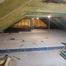 Poddasze z drewnianą konstrukcją dachu i deskami podłogowymi w trakcie montażu. Widoczne narzędzia i materiały budowlane. Oświetlenie punktowe.