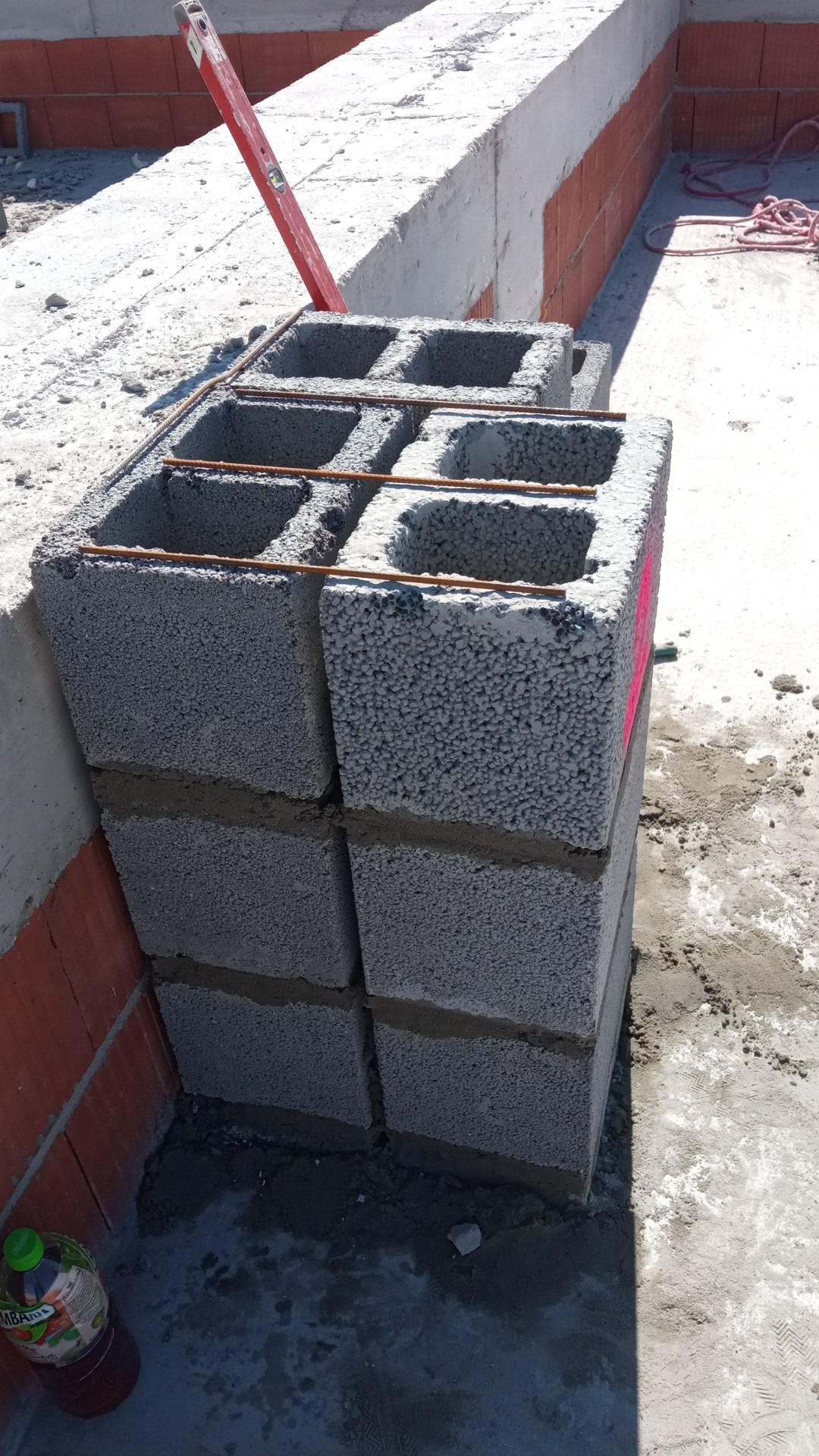 Wznoszenie ściany z szarych bloczków betonowych z widocznym zbrojeniem poziomym, poziomica oparta o ścianę z czerwonej cegły, butelka z napojem w rogu.