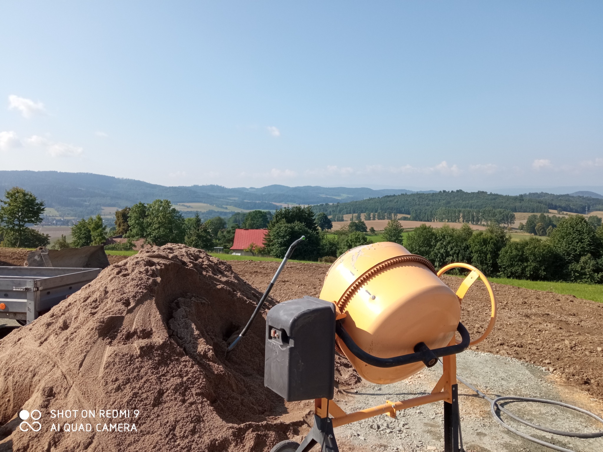 Żółta betoniarka stojąca obok pryzmy piasku na tle pagórkowatego krajobrazu z lasami i błękitnym niebem.