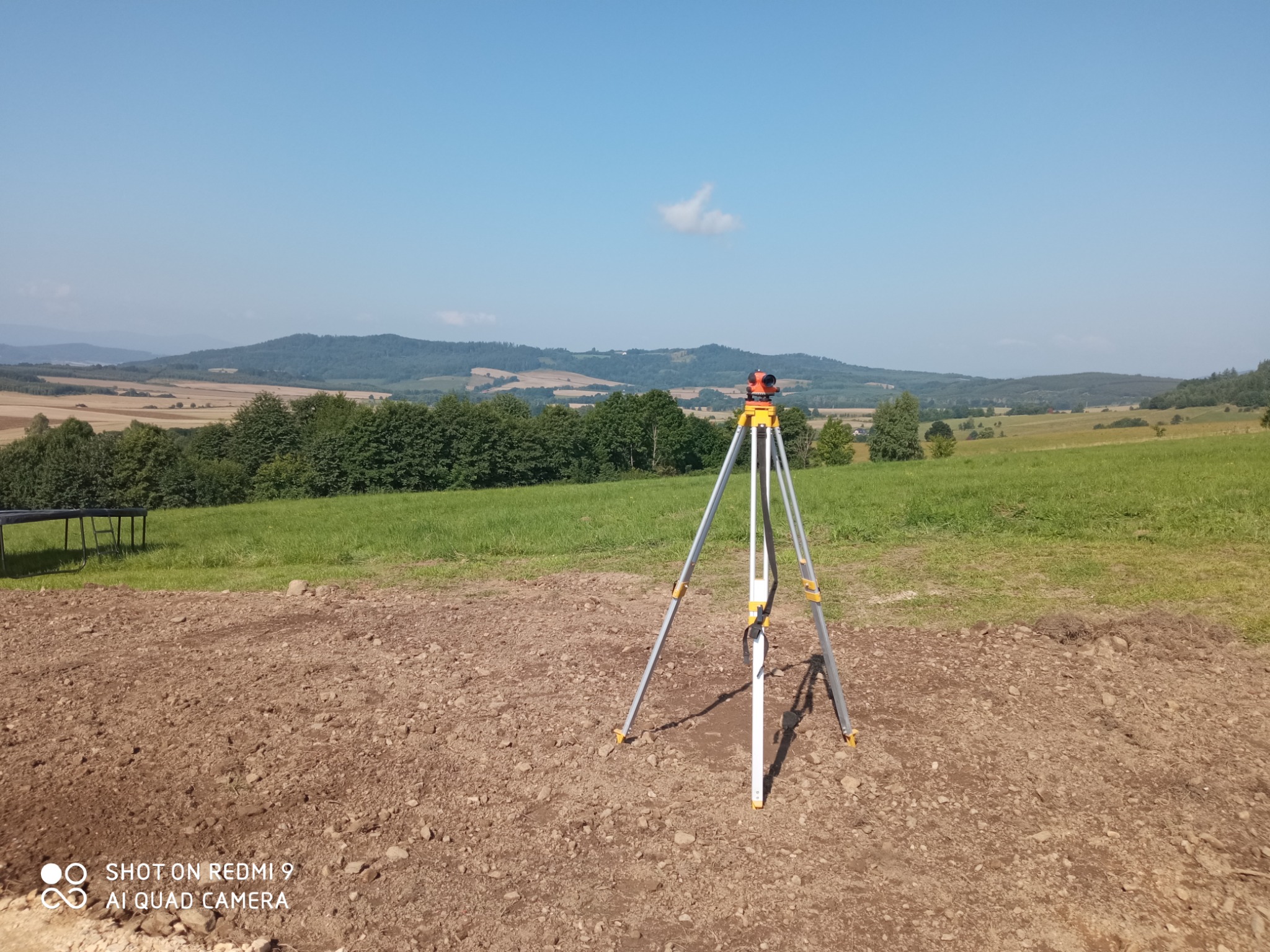 Geodezyjny niwelator optyczny ustawiony na trójnogu na przygotowanym pod budowę terenie, w tle zielone wzgórza i błękitne niebo z pojedynczą chmurą.