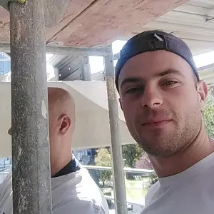 Portret młodego mężczyzny w czapce, uśmiechającego się do kamery na tle rusztowania i częściowo widocznej elewacji budynku w trakcie prac. W tle widoczny fragment innego pracownika i zabudowa miejska.