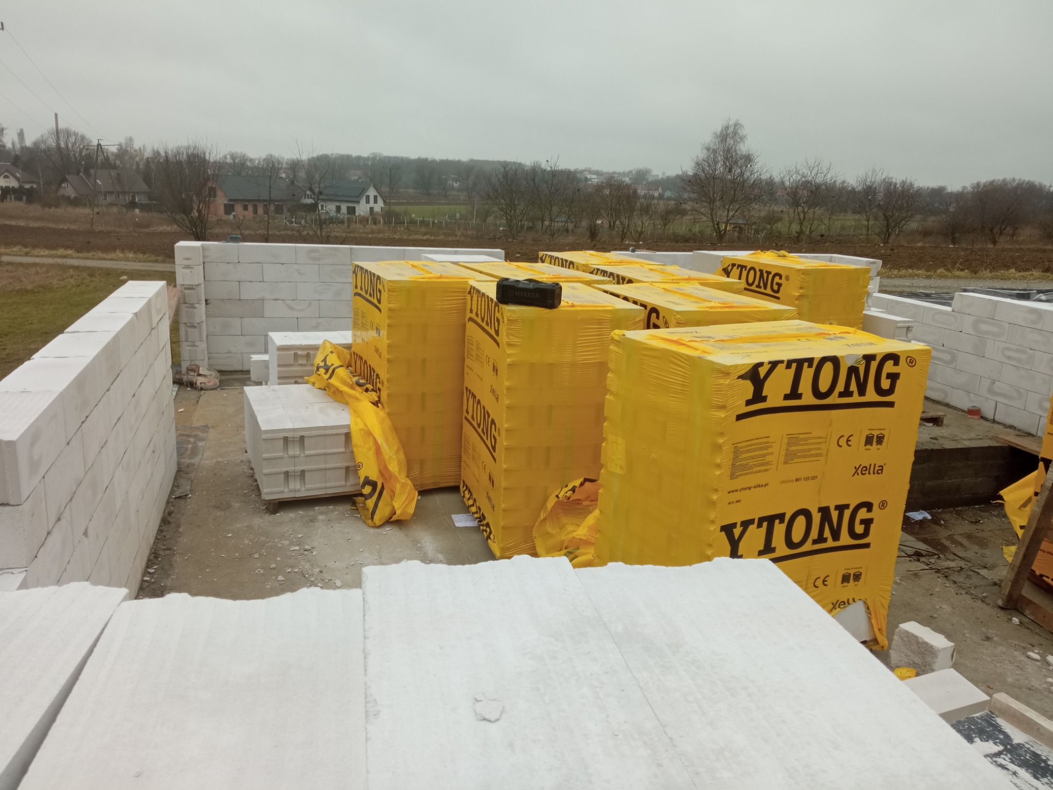 Budowa domu z bloczków Ytong, palety z zapakowanymi bloczkami na placu budowy, widoczne ściany w trakcie wznoszenia, krajobraz wiejski w tle.