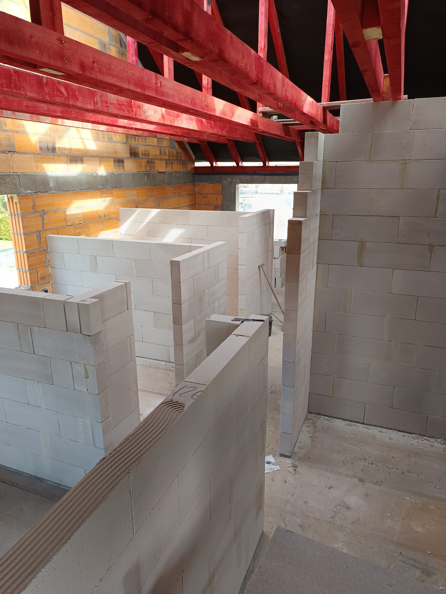 Wnętrze w trakcie budowy: ściany z białych bloczków, czerwona konstrukcja dachu, widoczne okno i fragment ściany z cegły. Na bloczkach widoczne ślady zaprawy.