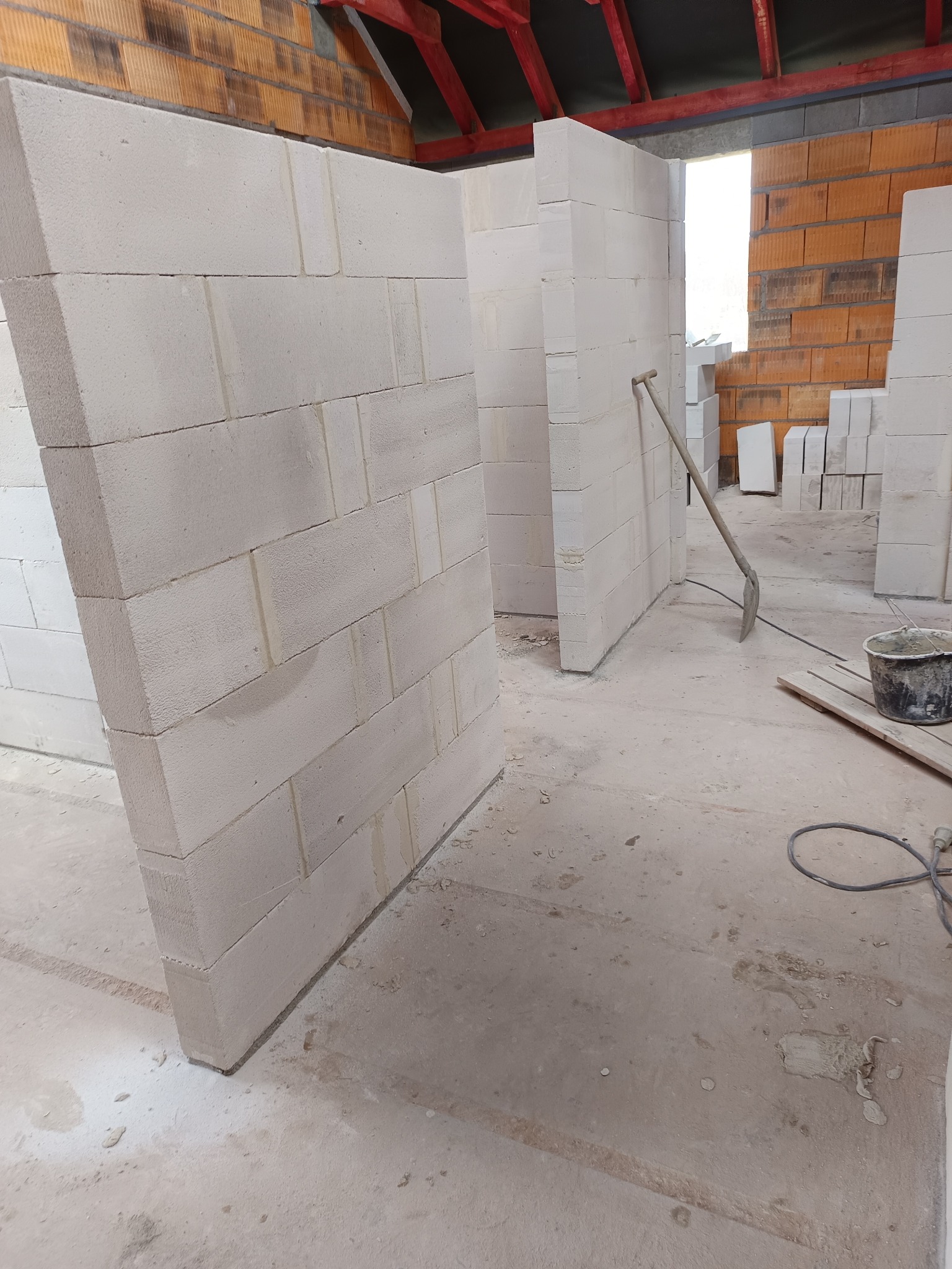 Wznoszenie ścian działowych z białych bloczków betonu komórkowego w surowym stanie, widoczny mur z cegły dziurawki w tle, narzędzia budowlane i wiadro z zaprawą.