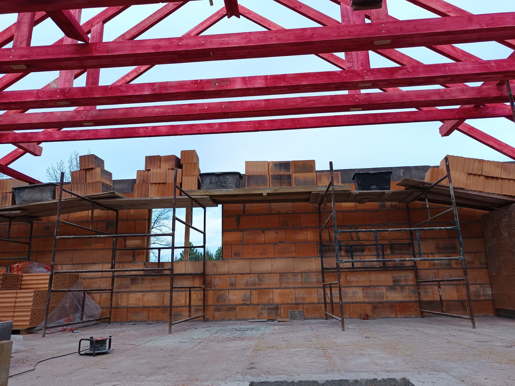 Wznoszenie ścian domu z czerwonej cegły ceramicznej, widoczne rusztowania i pomalowana na czerwono drewniana konstrukcja dachu.