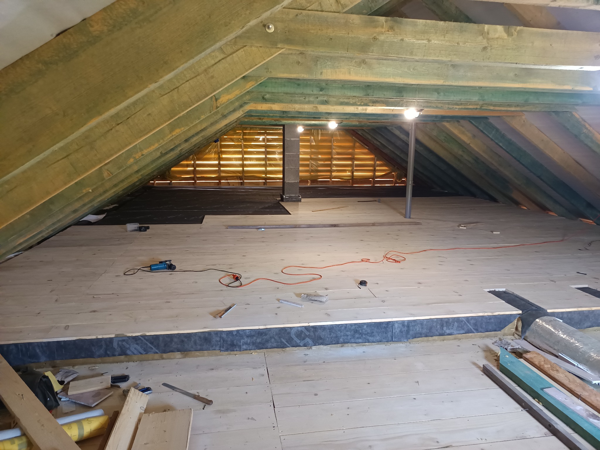 Poddasze z drewnianą konstrukcją dachu i deskami podłogowymi w trakcie montażu. Widoczne narzędzia i materiały budowlane. Oświetlenie punktowe.