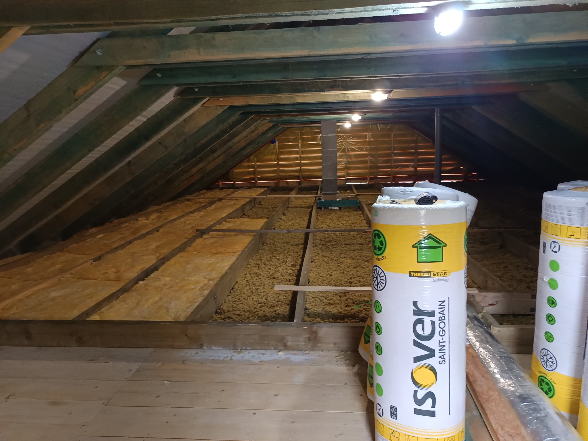 Poddasze z drewnianą konstrukcją i wełną mineralną Isover. Widoczne rolki materiału izolacyjnego. Oświetlenie sufitowe.