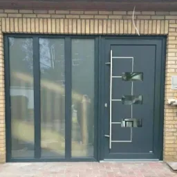 Szare drzwi zewnętrzne z trzema prostokątnymi przeszkleniami i białym, geometrycznym wzorem, połączone z trzema pionowymi oknami w czarnej ramie, osadzone w elewacji z żółtej cegły.