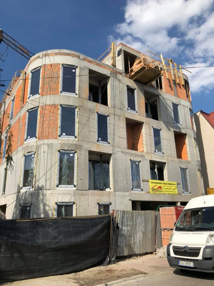 Budynek w trakcie budowy z zamontowanymi oknami w czarnych ramach, widoczny baner reklamowy firmy budowlanej i zaparkowany biały samochód dostawczy.