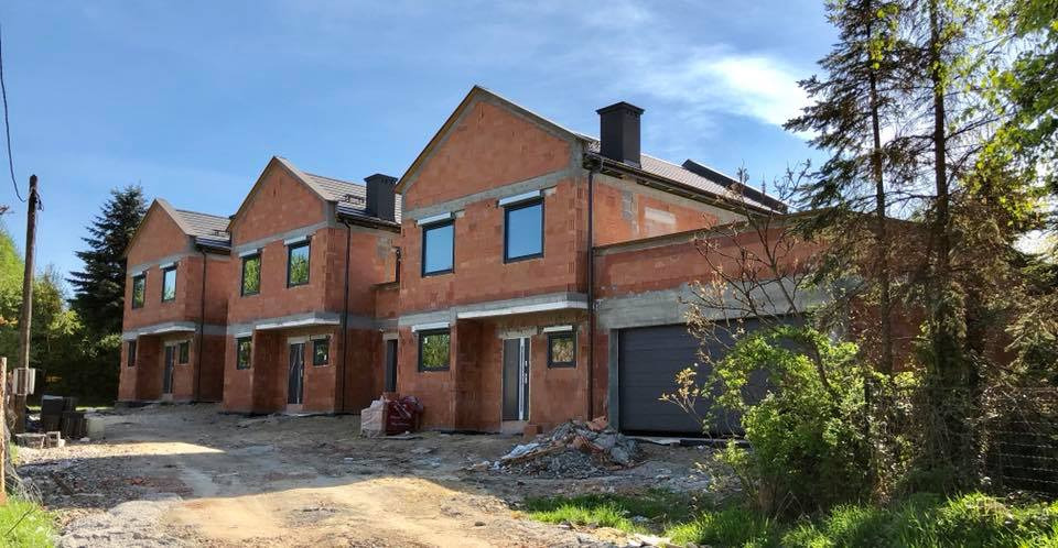 Surowe, szeregowe domy w trakcie budowy, z zamontowanymi oknami z czarnymi ramami i szarymi roletami zewnętrznymi, widoczne materiały budowlane i teren budowy.