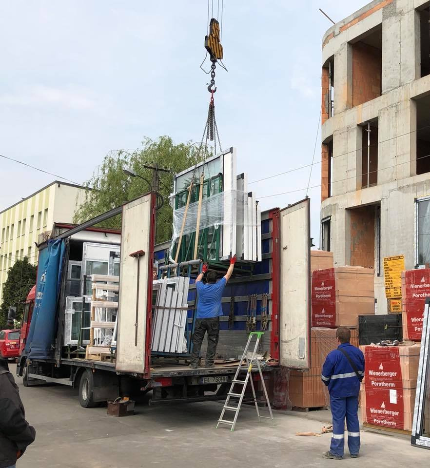 Rozładunek okien z ciężarówki za pomocą dźwigu na placu budowy, widoczny pracownik w czerwonych rękawicach i drabina oparta o skrzynię ładunkową.