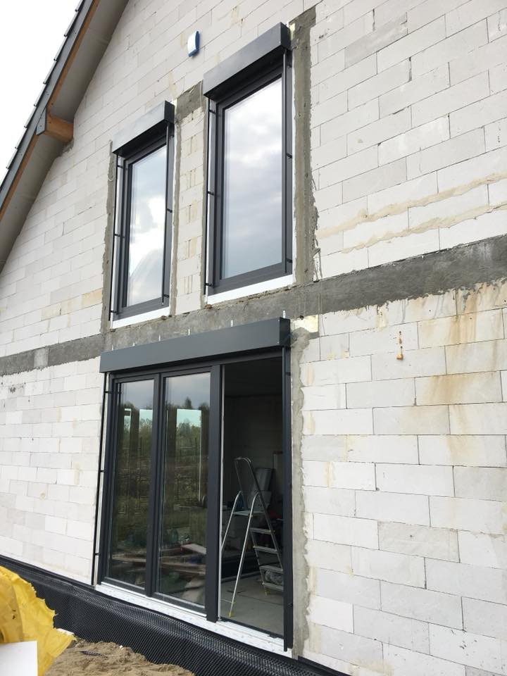 Wysoki budynek w trakcie budowy z zamontowanymi oknami PCV w czarnych ramach i roletami zewnętrznymi, widoczna drabina wewnątrz pomieszczenia przez otwarte okno tarasowe.