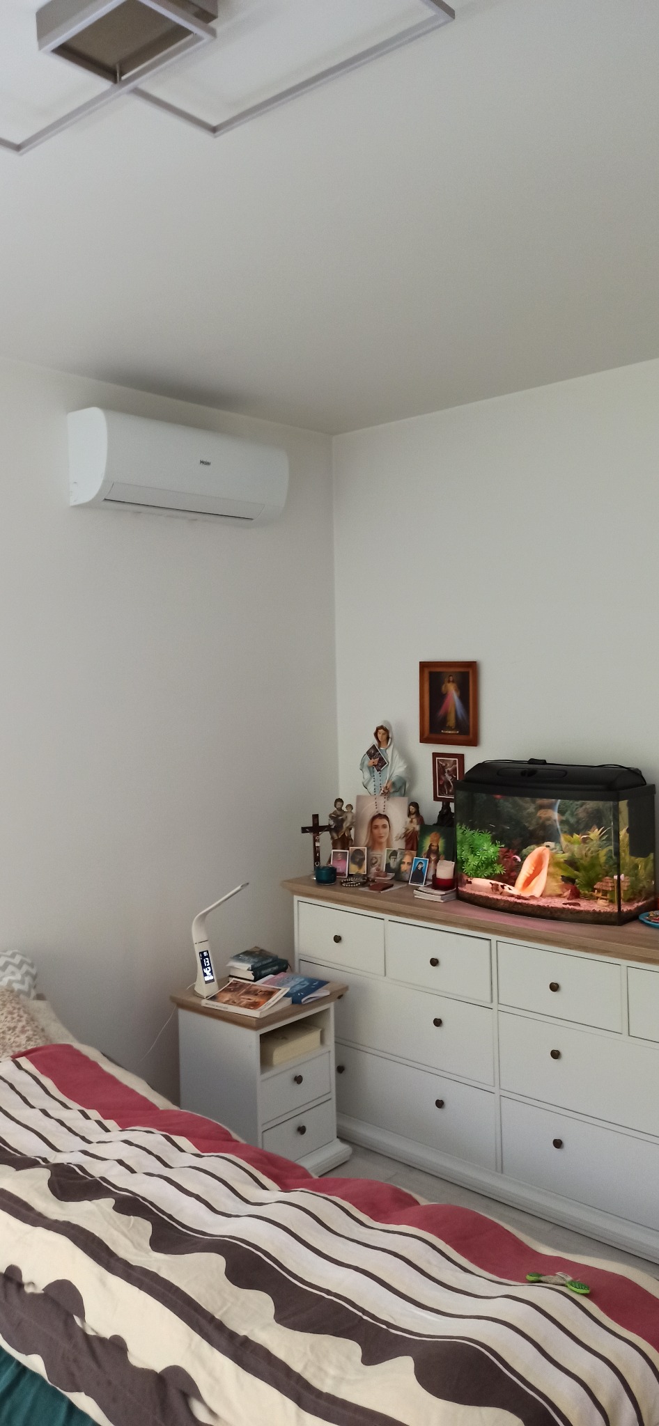Biały klimatyzator Haier zamontowany na ścianie w pokoju z białą komodą, akwarium i dekoracjami religijnymi