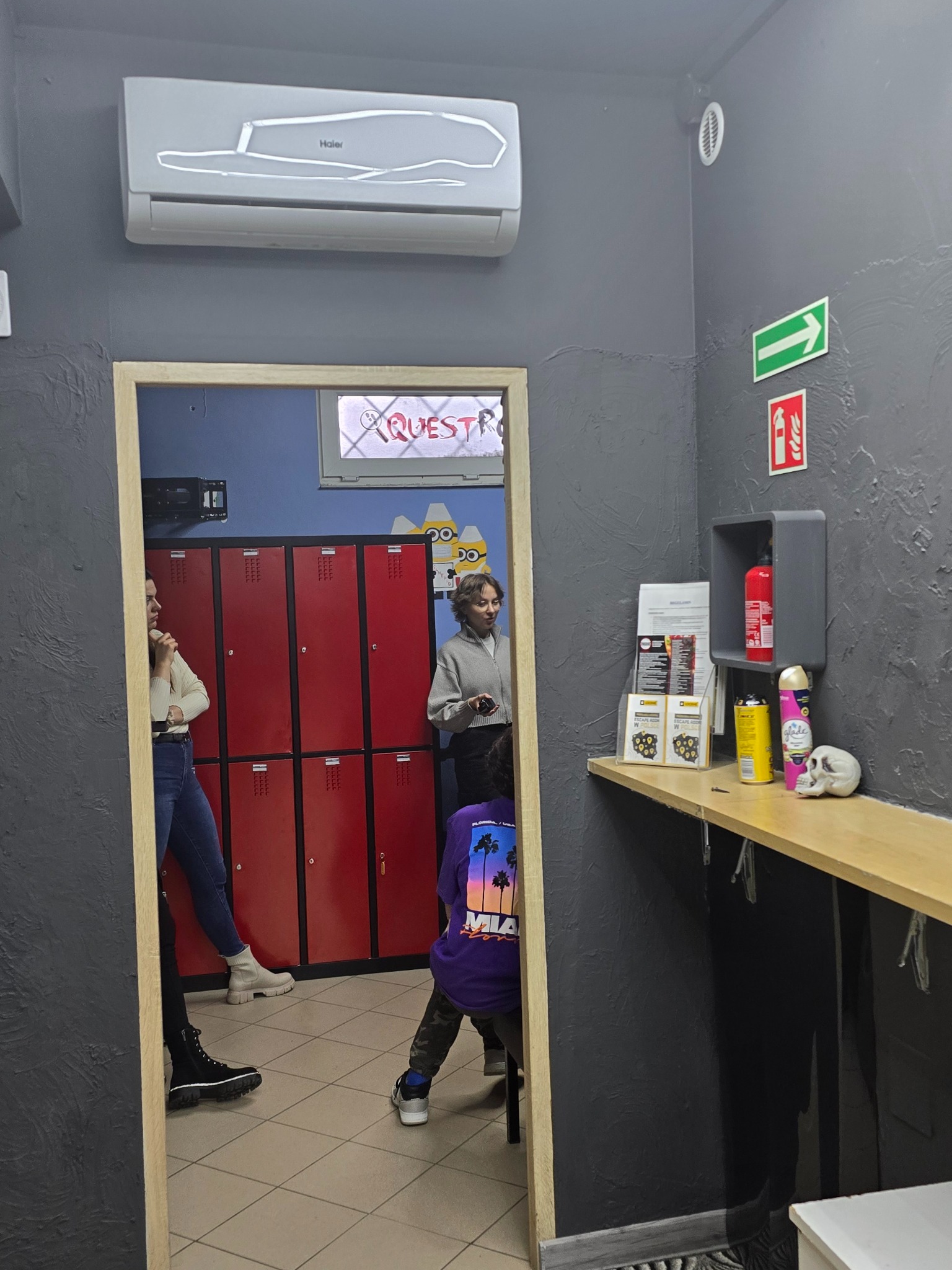 Biały klimatyzator Haier zamontowany na szarej ścianie w pomieszczeniu z czerwonymi szafkami, widoczny przez futrynę drzwiową, obok półki z rekwizytami escape roomu, takimi jak czaszka i spraye.