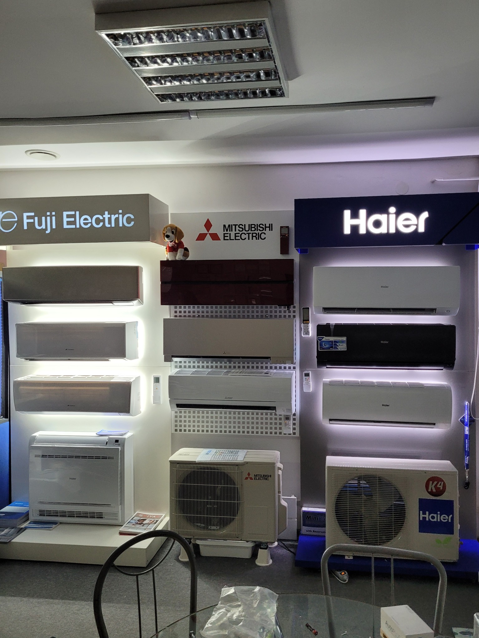 Ekspozycja klimatyzatorów różnych marek (Fuji Electric, Mitsubishi Electric, Haier) w salonie, z widocznymi jednostkami wewnętrznymi o zróżnicowanym designie i kolorystyce, oświetlonych intensywnym...