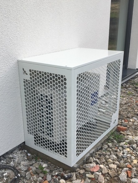 Zewnętrzna jednostka klimatyzacji zabezpieczona ażurową, białą obudową z motywem pszczoły, umieszczona przy ścianie budynku na żwirowym podłożu.