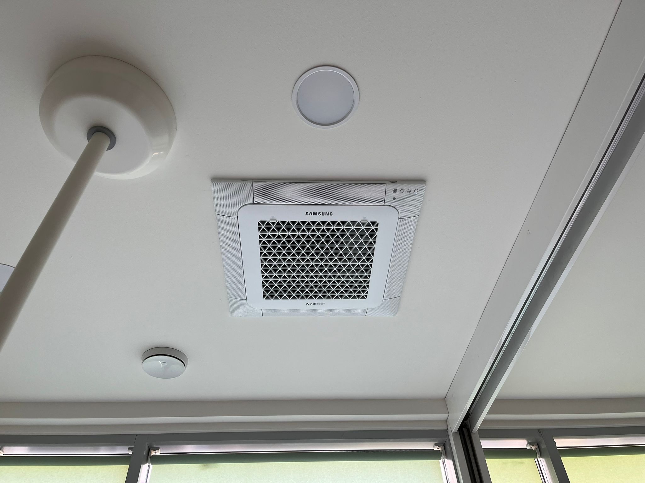 Kasetonowa jednostka klimatyzacji Samsung WindFree zamontowana na białym suficie biura, widoczne okna z roletami w tle.
