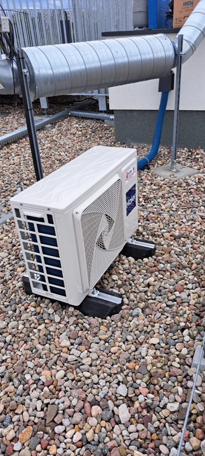 Biała jednostka zewnętrzna klimatyzacji Haier zamontowana na dachu pokrytym drobnymi kamieniami, z widocznymi przewodami i rurami wentylacyjnymi.