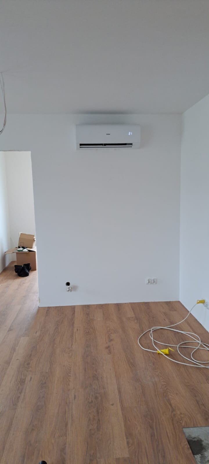 Świeżo zamontowany, biały klimatyzator Haier na białej ścianie w pokoju z panelami podłogowymi; widoczne gniazdka elektryczne i przewód z żółtą wtyczką na podłodze.