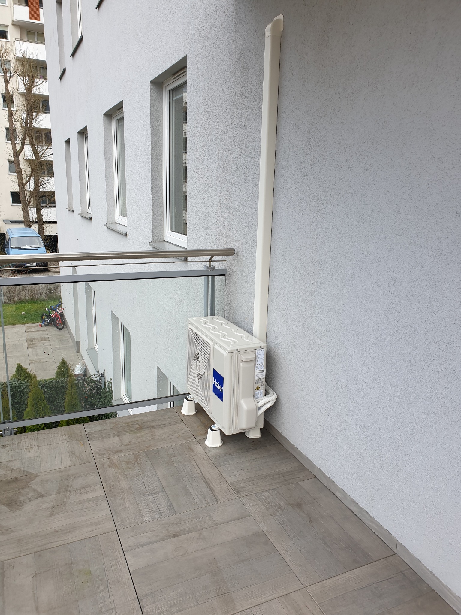 Biała jednostka zewnętrzna klimatyzacji Haier zamontowana na balkonie bloku mieszkalnego, z widoczną rurą odprowadzającą skropliny i osłoną instalacji biegnącą wzdłuż ściany.