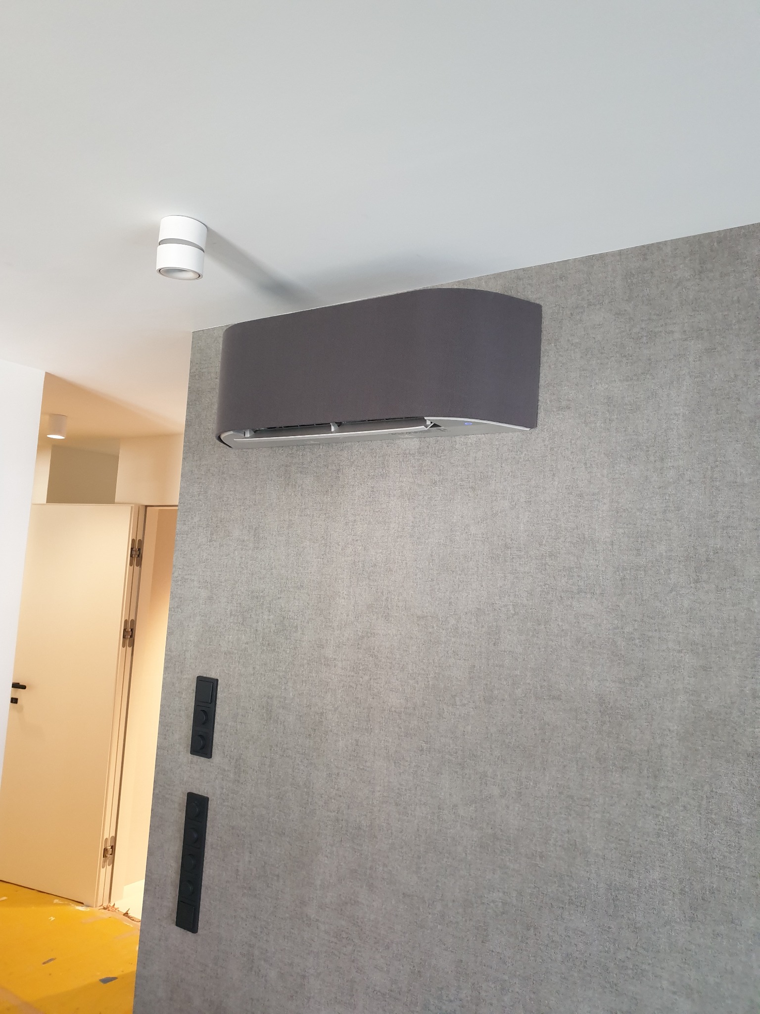 Szary klimatyzator ścienny o zaokrąglonym kształcie w nowoczesnym wnętrzu z szarą tapetą i czarnymi włącznikami, widoczny fragment białych drzwi i żółta folia ochronna na podłodze.