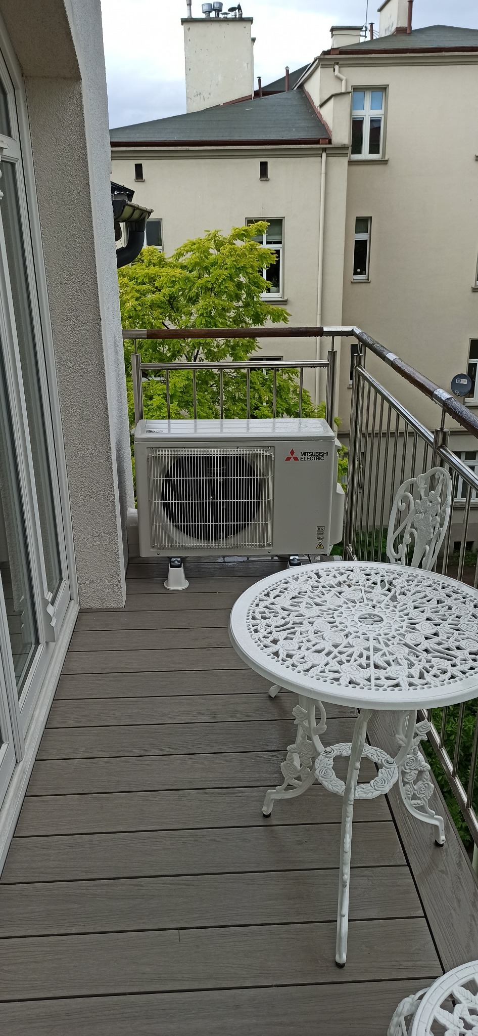 Zewnętrzna jednostka klimatyzacji Mitsubishi Electric zamontowana na balkonie z drewnianą podłogą, obok ażurowego, białego stolika i krzesła, widok na budynek mieszkalny i drzewa.