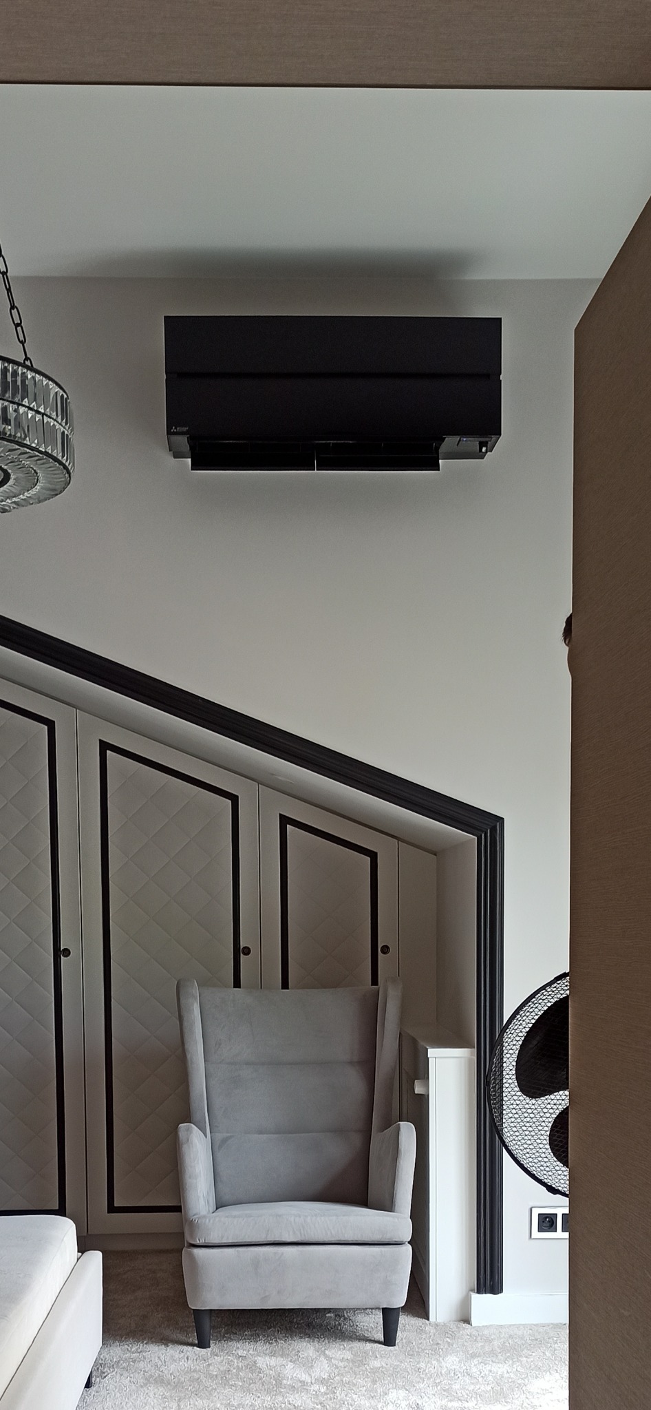 Czarna jednostka klimatyzacji zamontowana na ścianie w nowoczesnym wnętrzu nad szarym fotelem uszakiem, widoczna część kryształowego żyrandola i zabudowa szafy z dekoracyjnymi panelami.