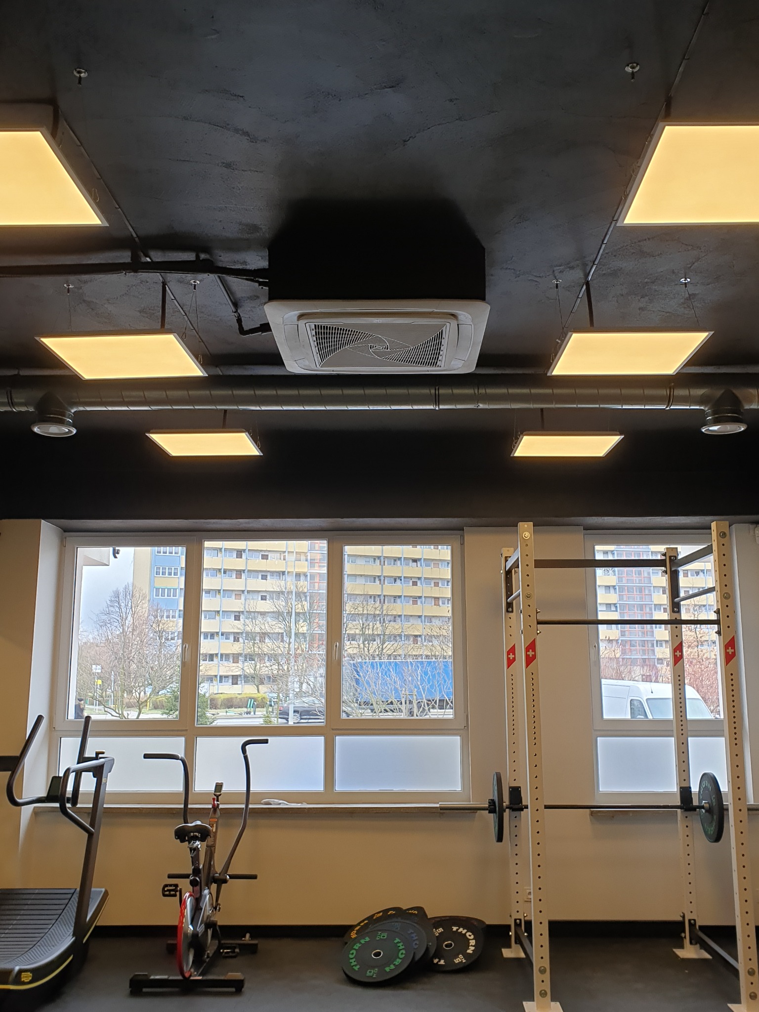Sufit w siłowni z zamontowaną kasetą klimatyzacji centralnej i kwadratowymi panelami LED, widok na okno z widokiem na budynki mieszkalne.