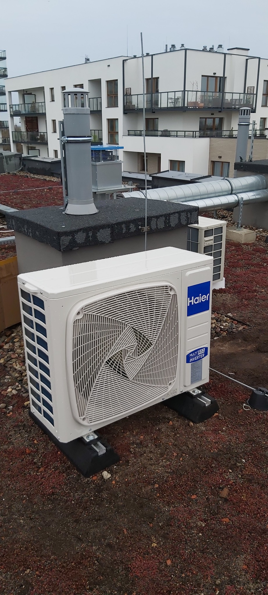 Zewnętrzna jednostka klimatyzacji Haier zamontowana na dachu budynku mieszkalnego z widocznymi elementami instalacji wentylacyjnej w tle.