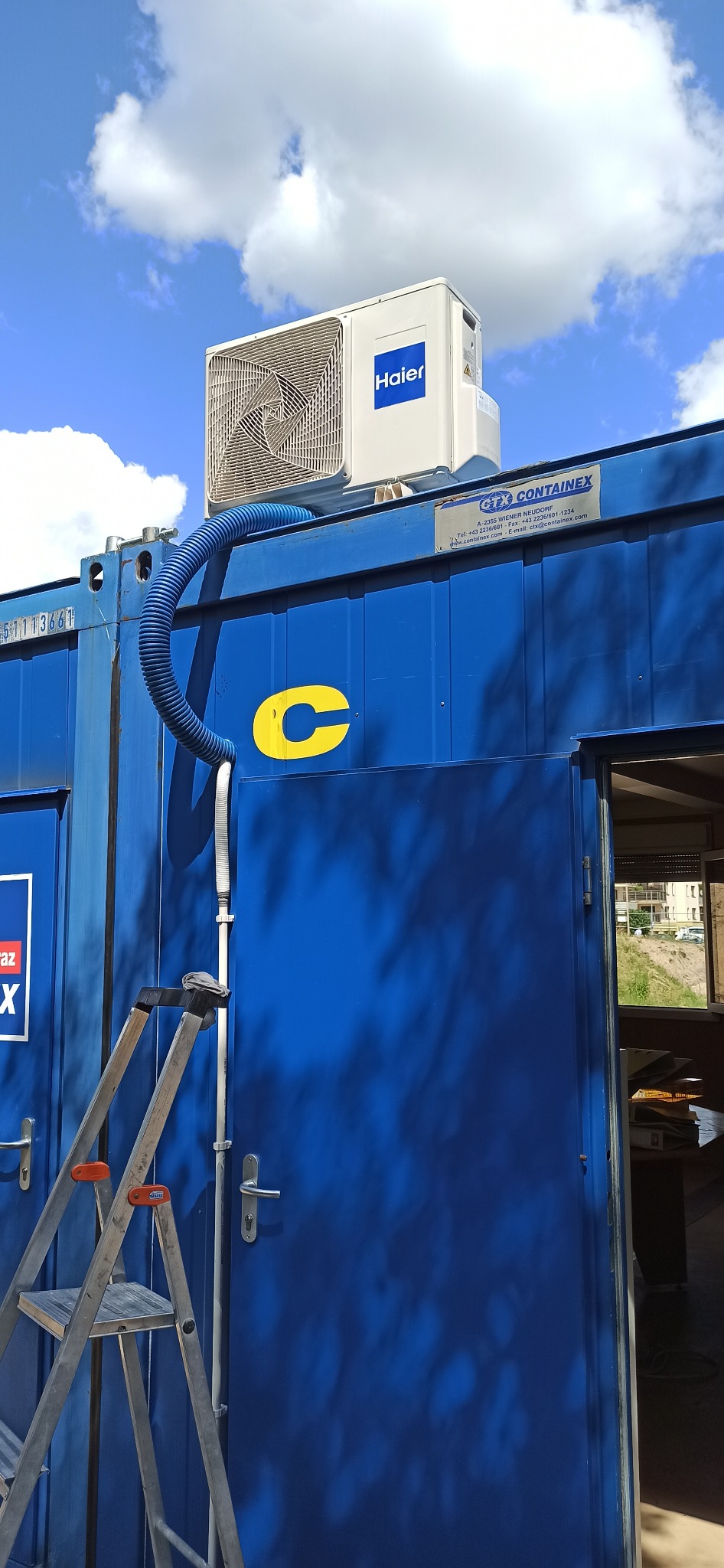 Klimatyzator Haier zamontowany na niebieskim kontenerze biurowym z widoczną drabiną i niebieskim przewodem wentylacyjnym na tle błękitnego nieba.