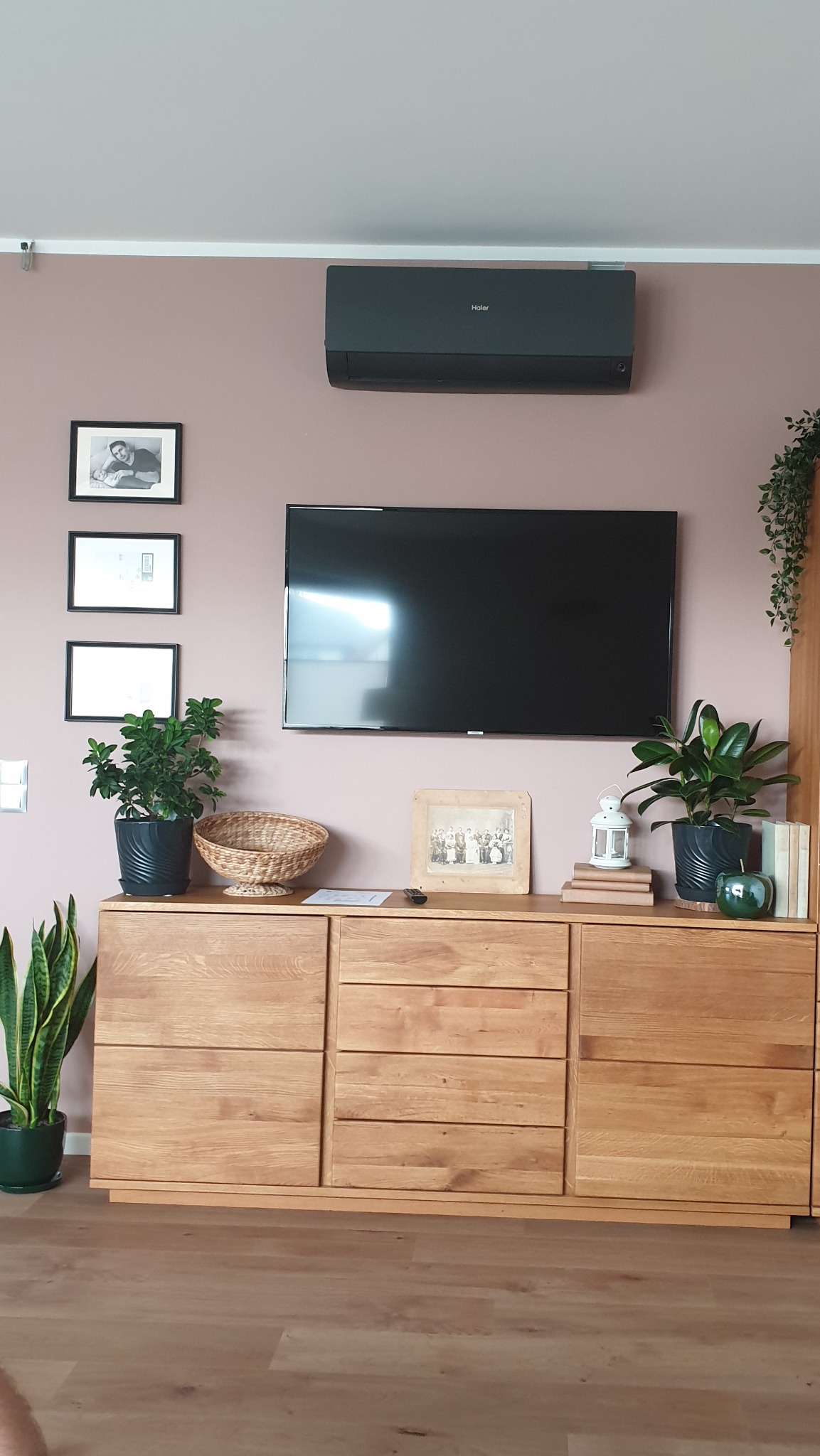 Nowoczesny salon z szarą klimatyzacją Haier zamontowaną nad telewizorem, drewniana komoda z dekoracjami i roślinami, ściana w kolorze pudrowego różu.