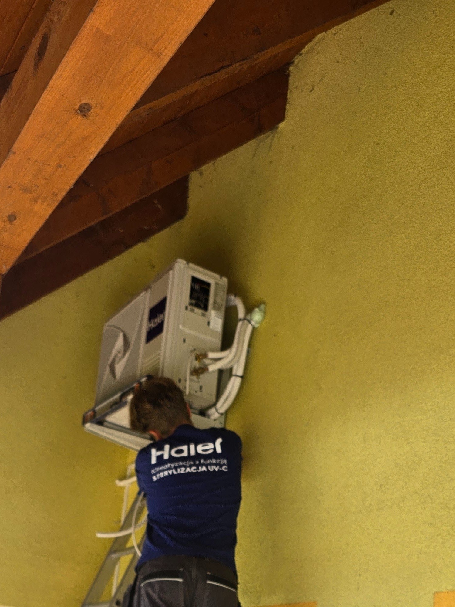Montaż klimatyzacji Haier na poddaszu, widoczny pracownik na drabinie podłączający przewody, drewniane belki stropowe i żółta ściana.