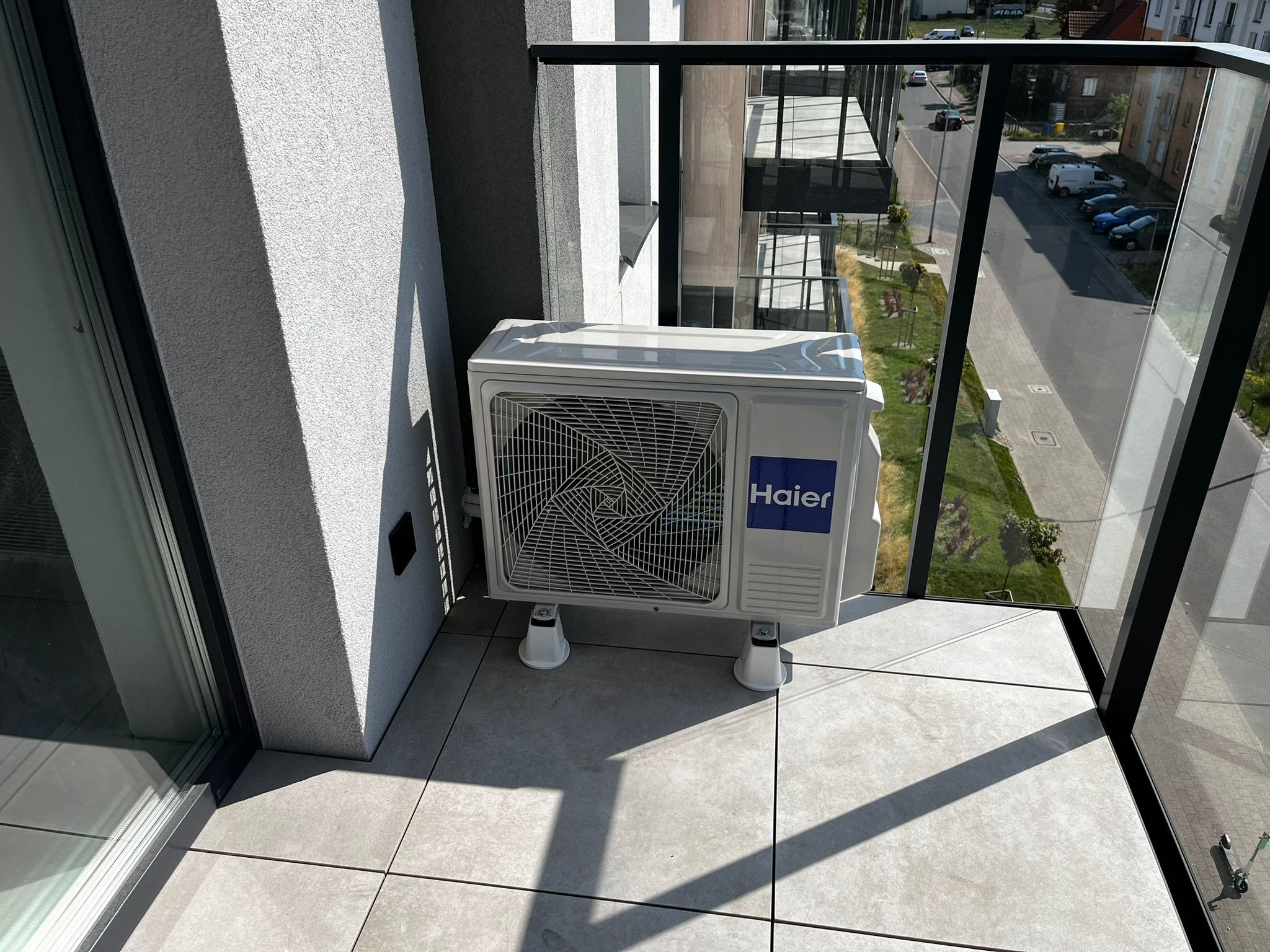 Biała jednostka zewnętrzna klimatyzacji Haier zamontowana na balkonie z szarymi płytkami, widok na ulicę przez szklaną balustradę.