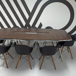 Nowoczesna sala konferencyjna z dużym, wielokątnym stołem z wbudowanymi gniazdami i ergonomicznymi krzesłami na drewnianych nogach, na tle ściany z geometrycznym wzorem.