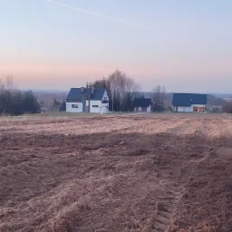 Ciężki traktor z podłączonym sprzętem rolniczym na świeżo zaoranym polu, w tle zabudowania mieszkalne i drzewa oświetlone zachodzącym słońcem.