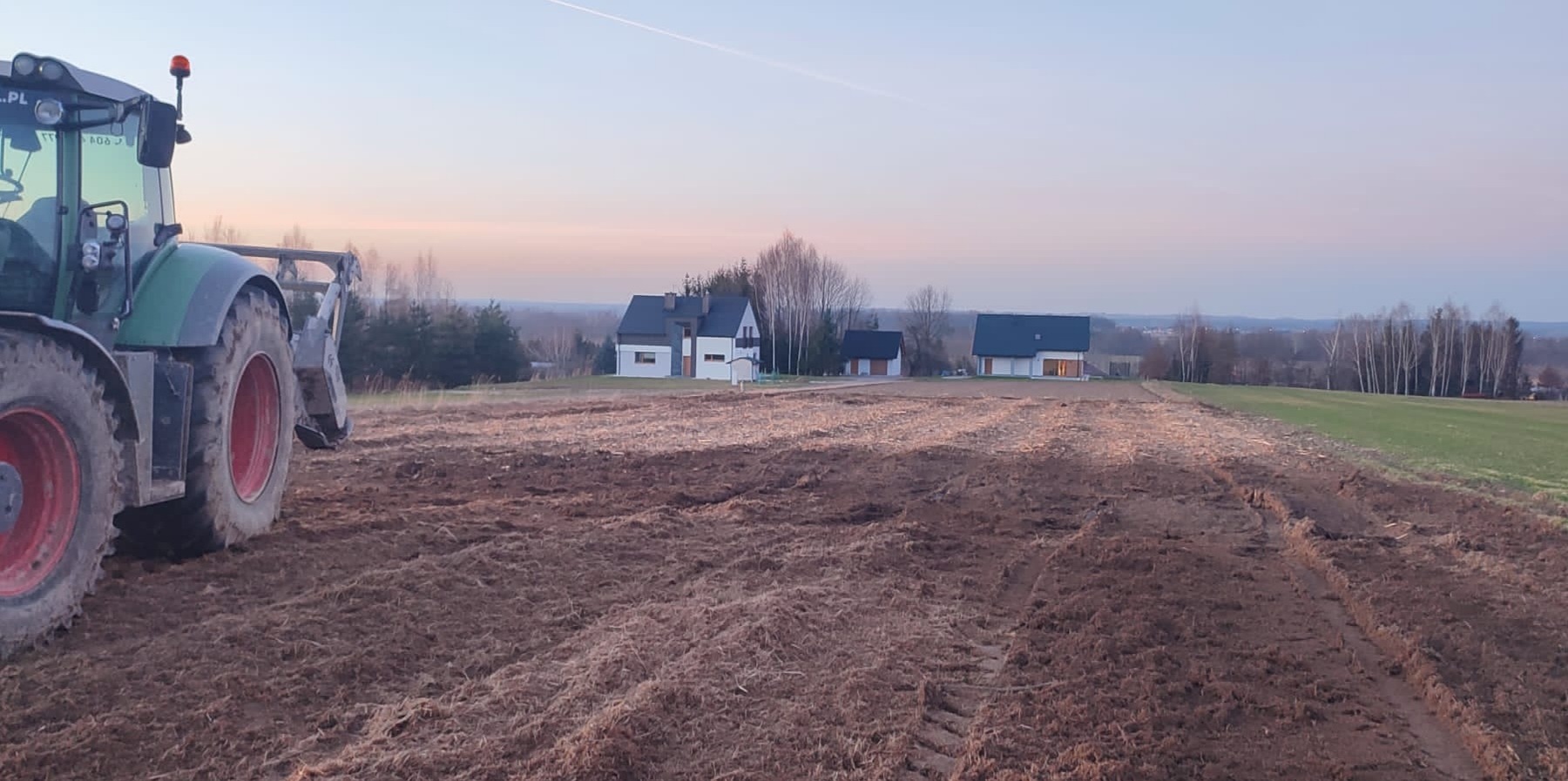 Ciężki traktor z podłączonym sprzętem rolniczym na świeżo zaoranym polu, w tle zabudowania mieszkalne i drzewa oświetlone zachodzącym słońcem.