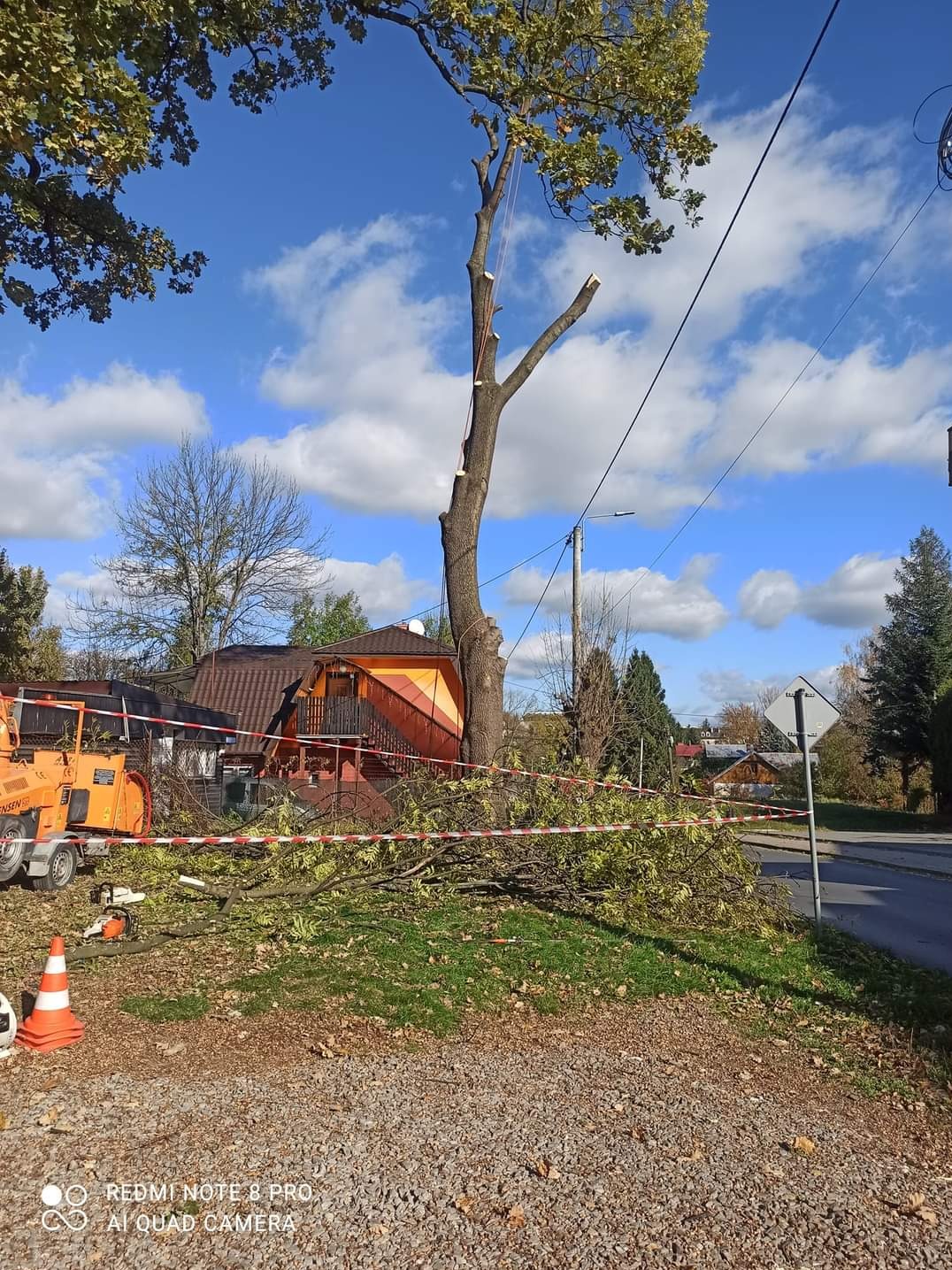 Częściowo ogołocone drzewo z linami zabezpieczającymi, obok rozdrabniacz gałęzi i piła łańcuchowa, teren zabezpieczony taśmą, w tle dom z pomarańczową elewacją i błękitne niebo z chmurami.