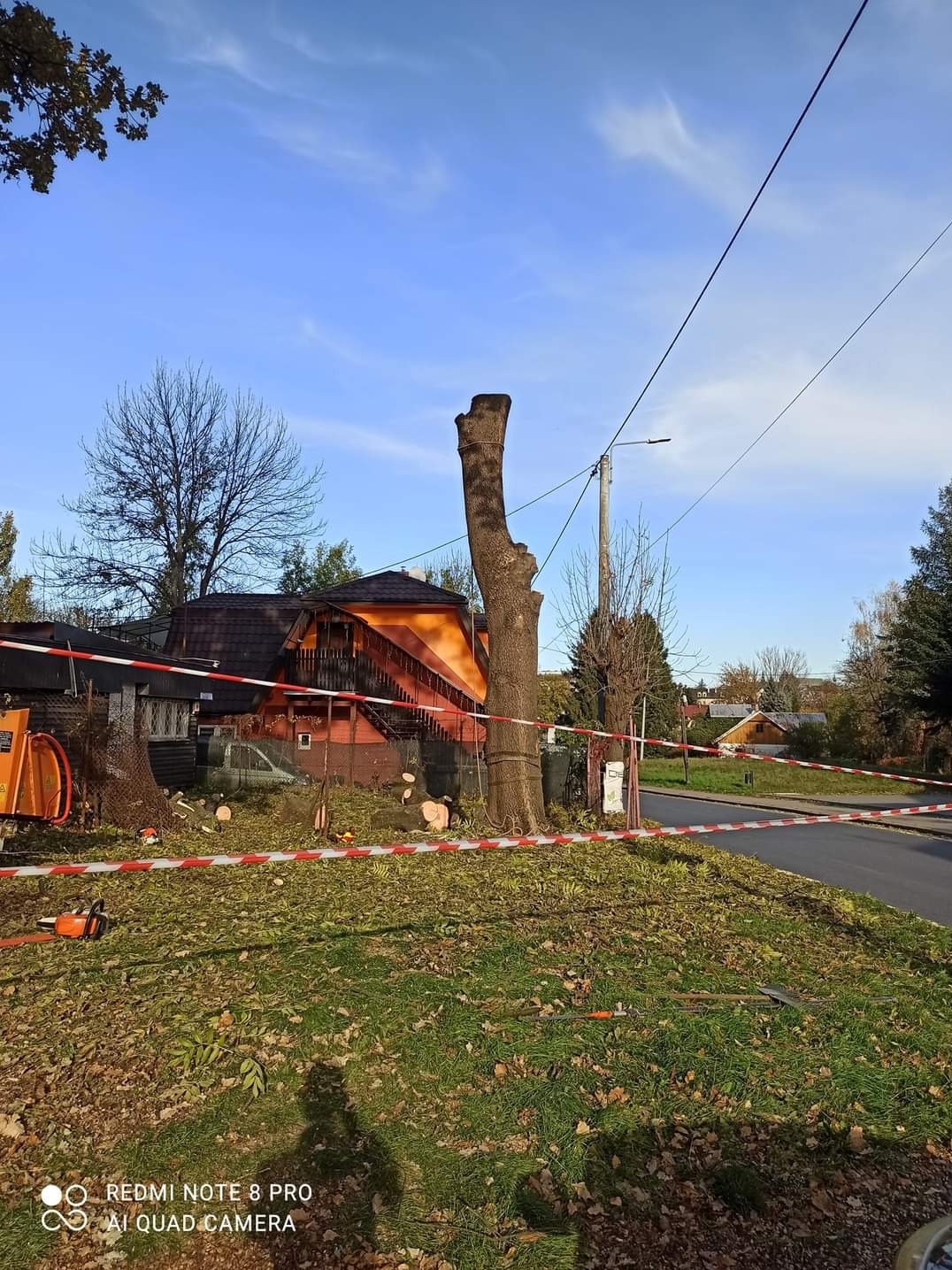 Przycięte drzewo zabezpieczone taśmą ostrzegawczą, w tle dom z pomarańczową elewacją i fragment maszyny do cięcia drewna.