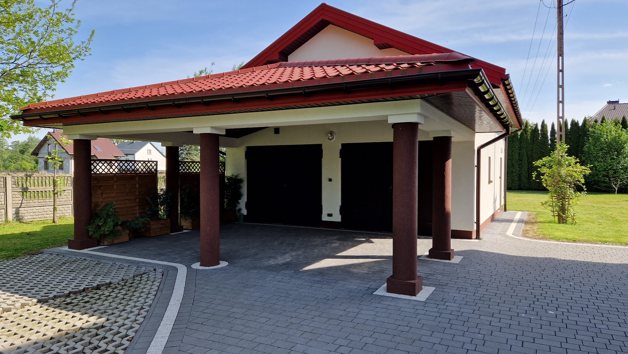 Zadaszone miejsce postojowe dla dwóch samochodów z czerwonym dachem i brązowymi kolumnami, wyłożone szarą kostką brukową.