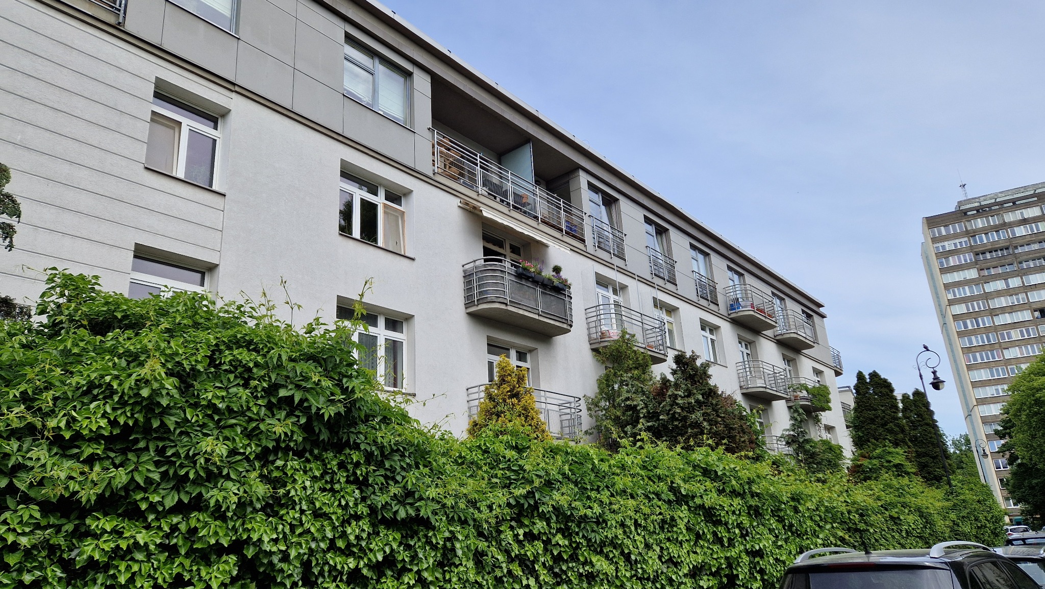 Wielopiętrowy budynek mieszkalny z balkonami, częściowo zasłonięty zielonym bluszczem i drzewami, z widocznym w tle wieżowcem.