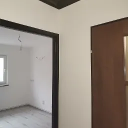 Wnętrze po remoncie: białe ściany, szara podłoga z paneli, czarne obramowania drzwi i sufitu, widoczne gniazdka elektryczne i przewody wystające ze ściany w pokoju obok.