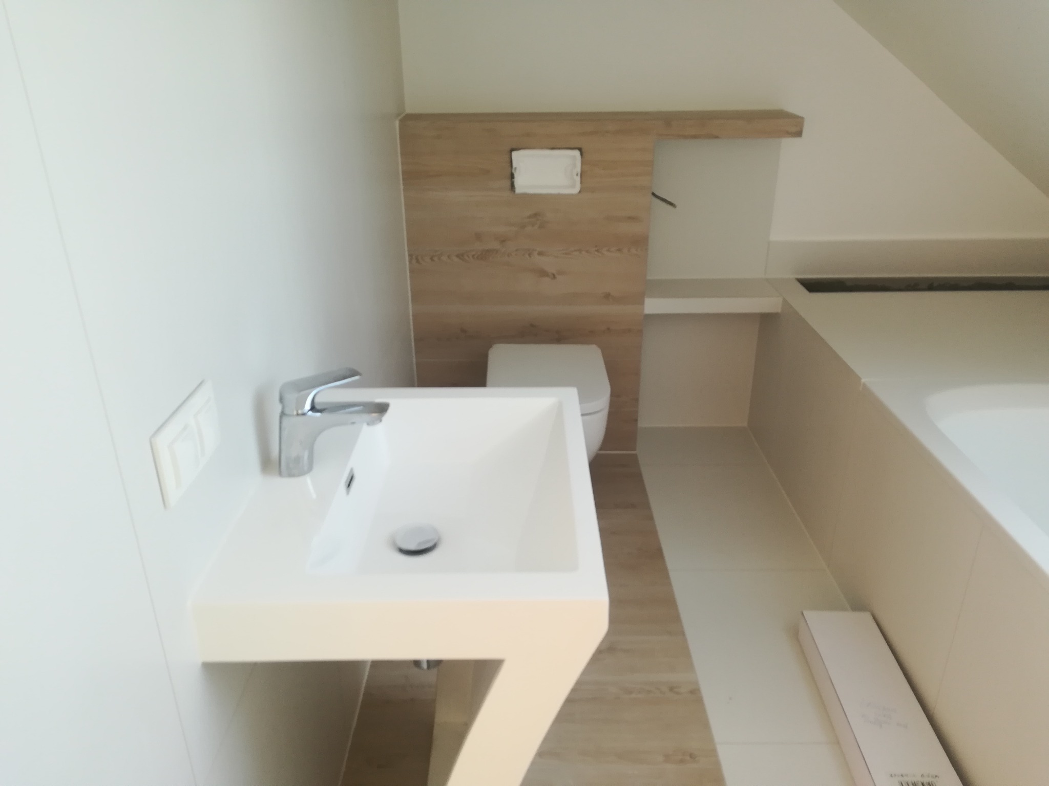 Nowoczesna, jasna łazienka z białą umywalką na postumencie, toaletą w zabudowie z drewnopodobną okładziną i fragmentem wanny; widoczne włączniki światła na ścianie.