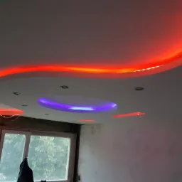 Sufit podwieszany z oświetleniem LED: czerwone światło obwodowe i centralne, okrągłe światło fioletowe, widok na okno z zielenią na zewnątrz, ściana częściowo wykończona.