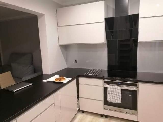 Nowoczesna kuchnia w Słupsku z czarnym blatem, białymi szafkami i piekarnikiem z ręcznikiem, widok na salon.