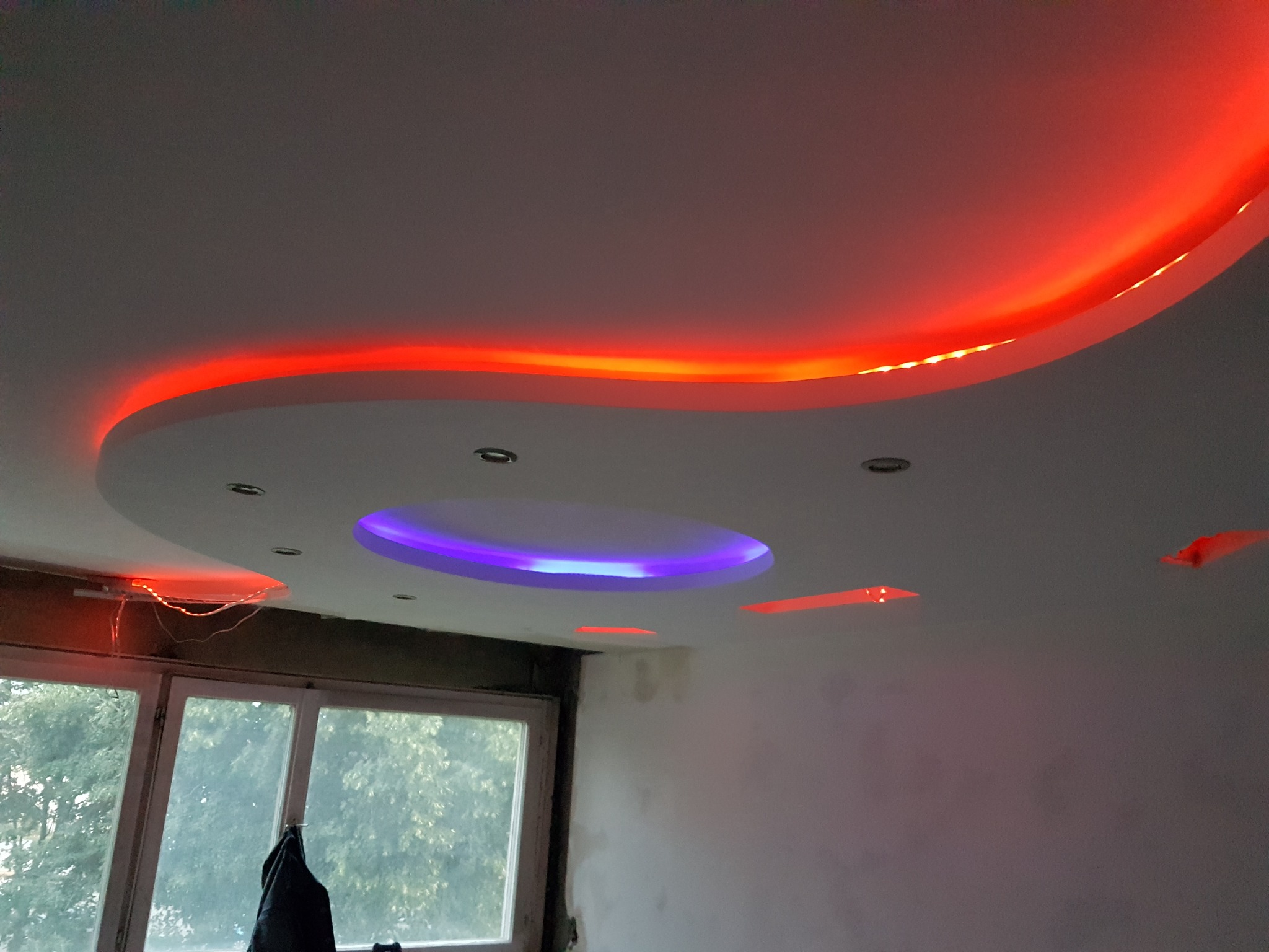 Sufit podwieszany z oświetleniem LED: czerwone światło obwodowe i centralne, okrągłe światło fioletowe, widok na okno z zielenią na zewnątrz, ściana częściowo wykończona.